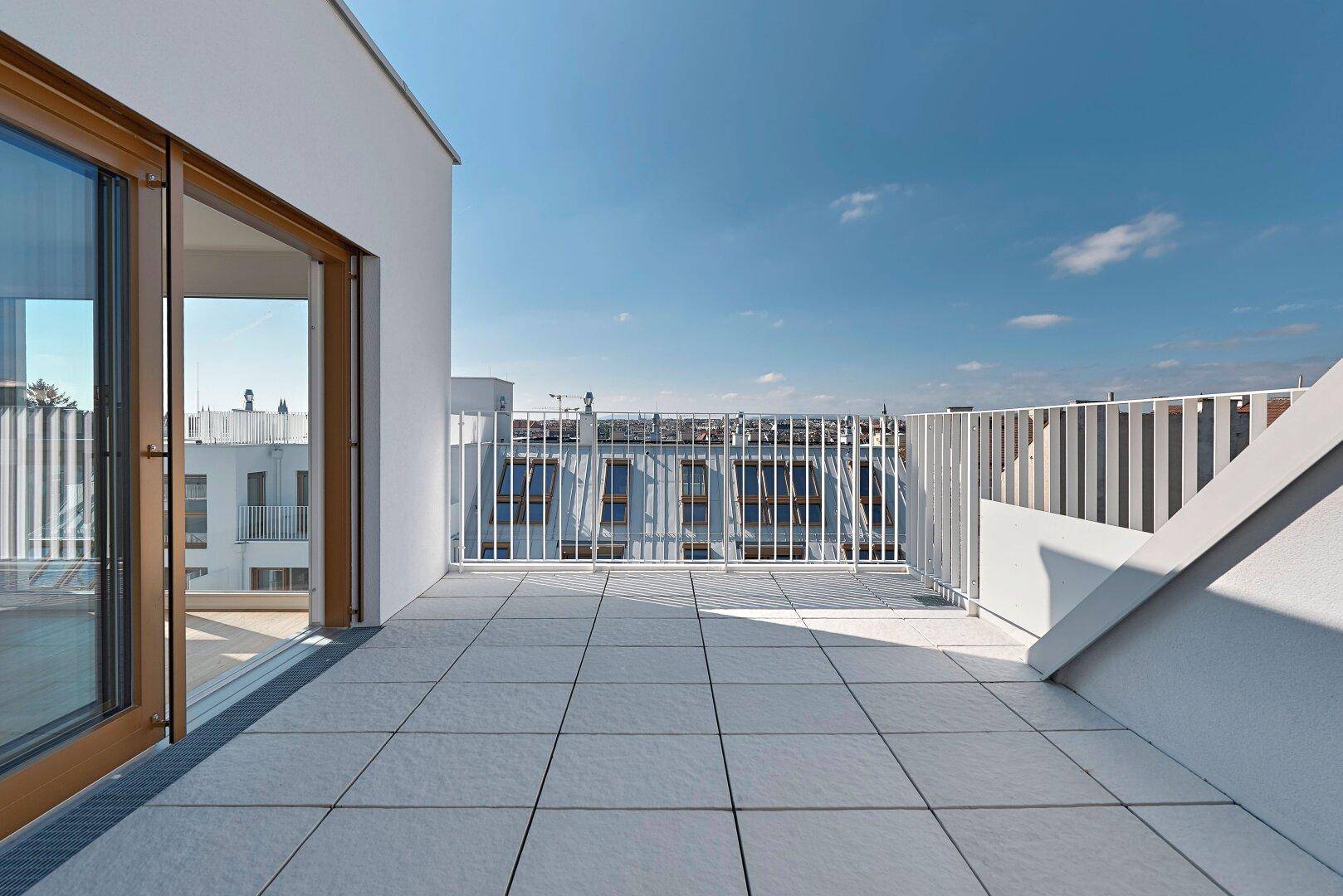 Weitläufige Dachterrasse mit Fliesenboden, weißem Geländer und Blick auf die Stadt.