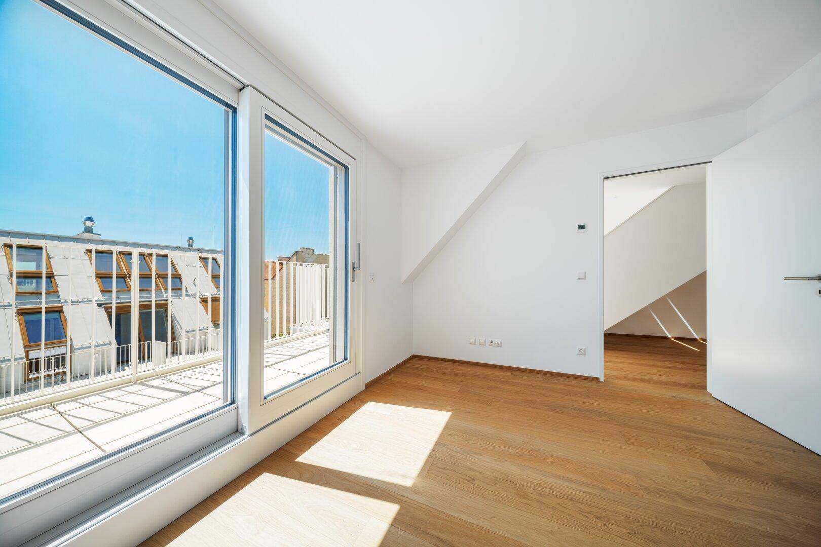 Helles Zimmer mit Schiebetür zur Terrasse, Holzboden und Blick auf umliegende Gebäude.