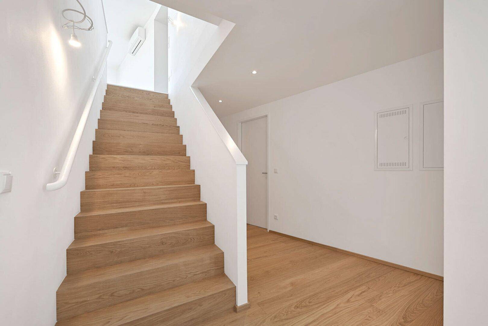 Moderne Holztreppe mit weißem Geländer, die zu einem hellen oberen Stockwerk des Gebäudes führt.