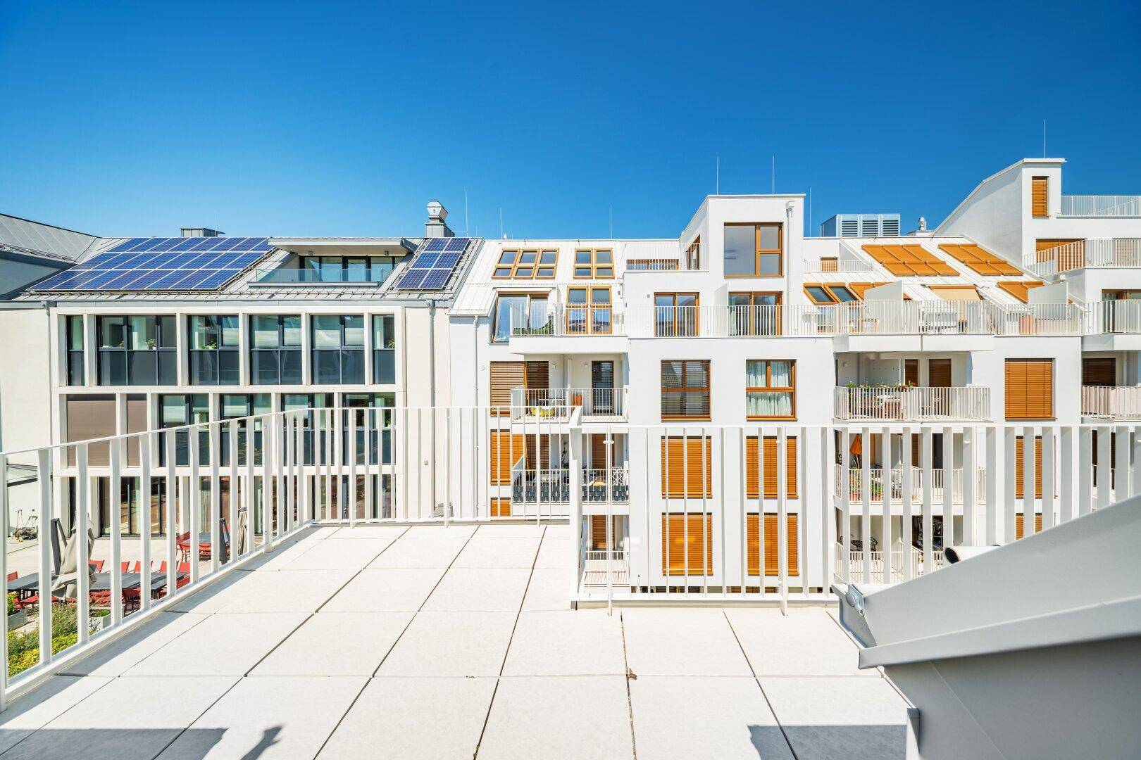 Moderne weiße Wohnanlage mit zahlreichen Balkonen, großen Fenstern und Solarpaneelen auf dem Dach.