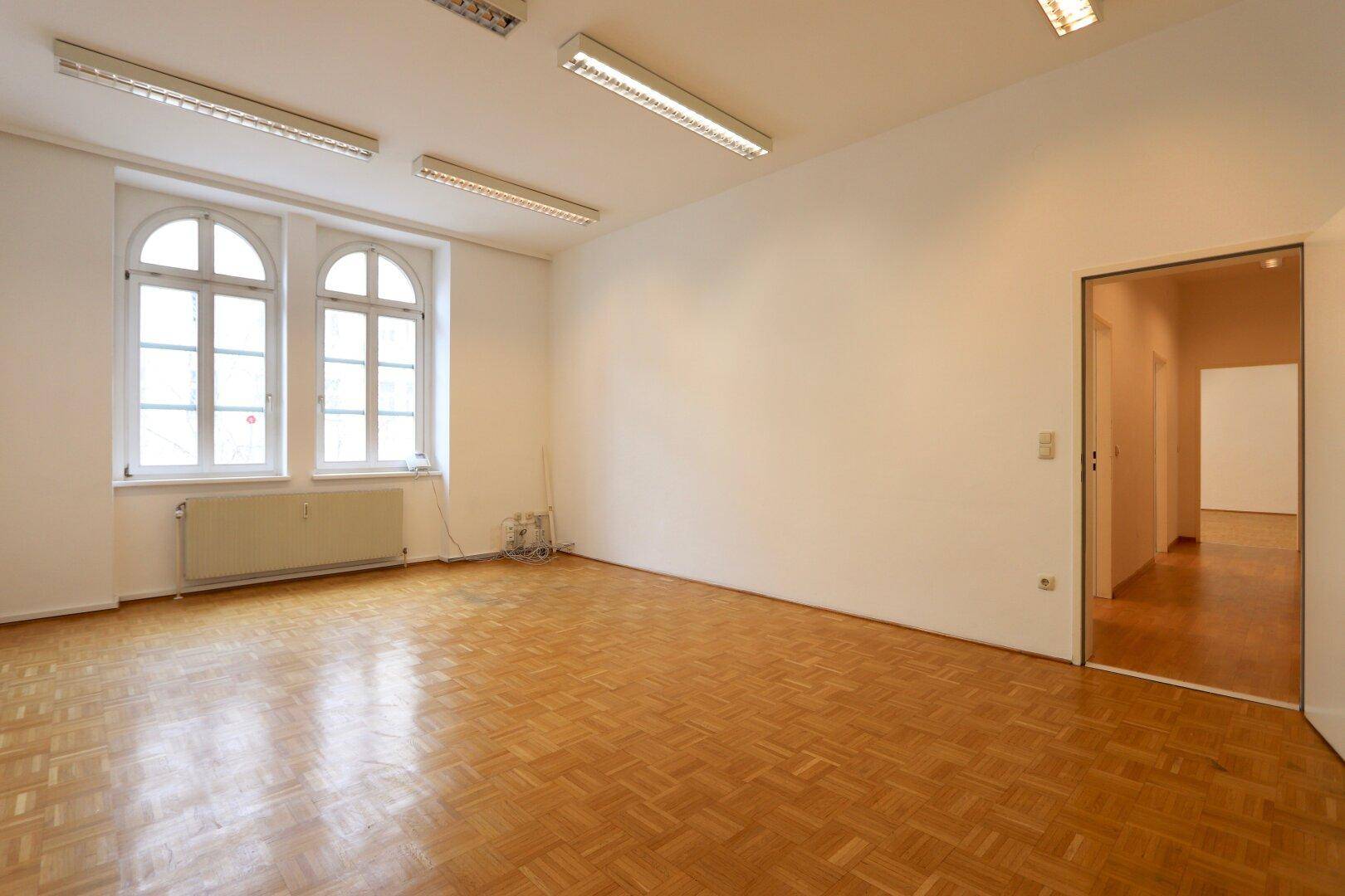 Heller, leerer Raum mit Fischgrätparkett, hohen Fenstern und funktionaler Deckenbeleuchtung.
