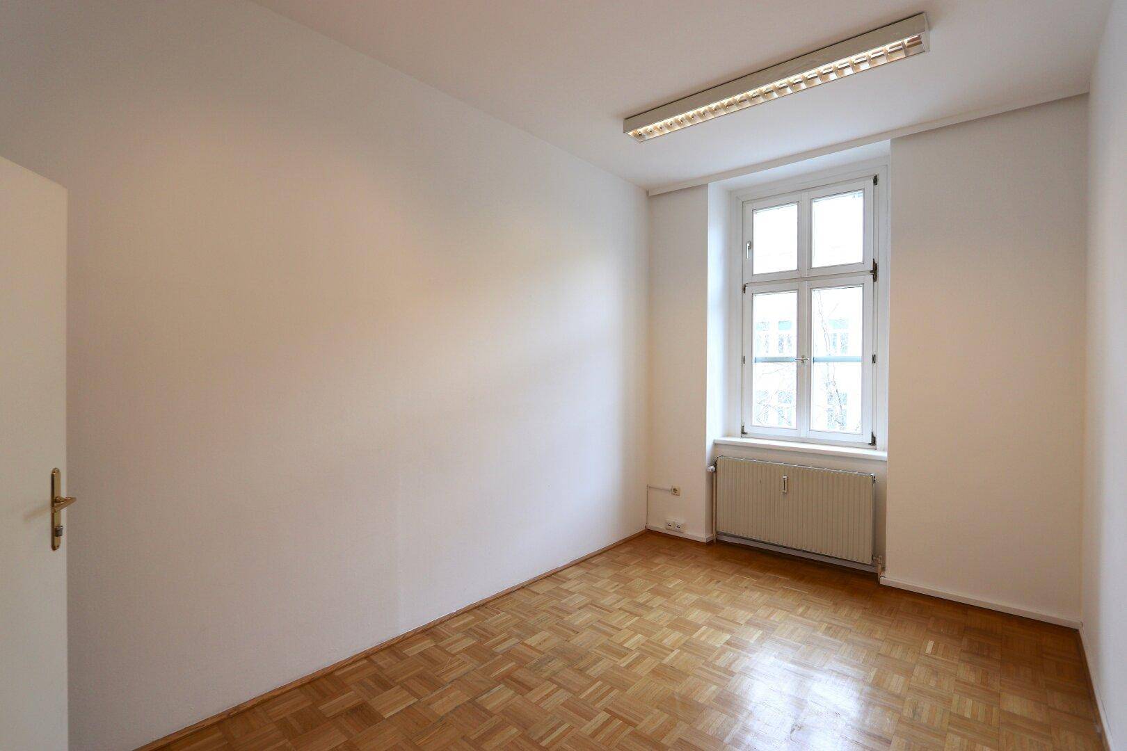 Kleinerer, heller Raum mit Parkettboden, einem Fenster und Heizkörper, ideal als Büro.