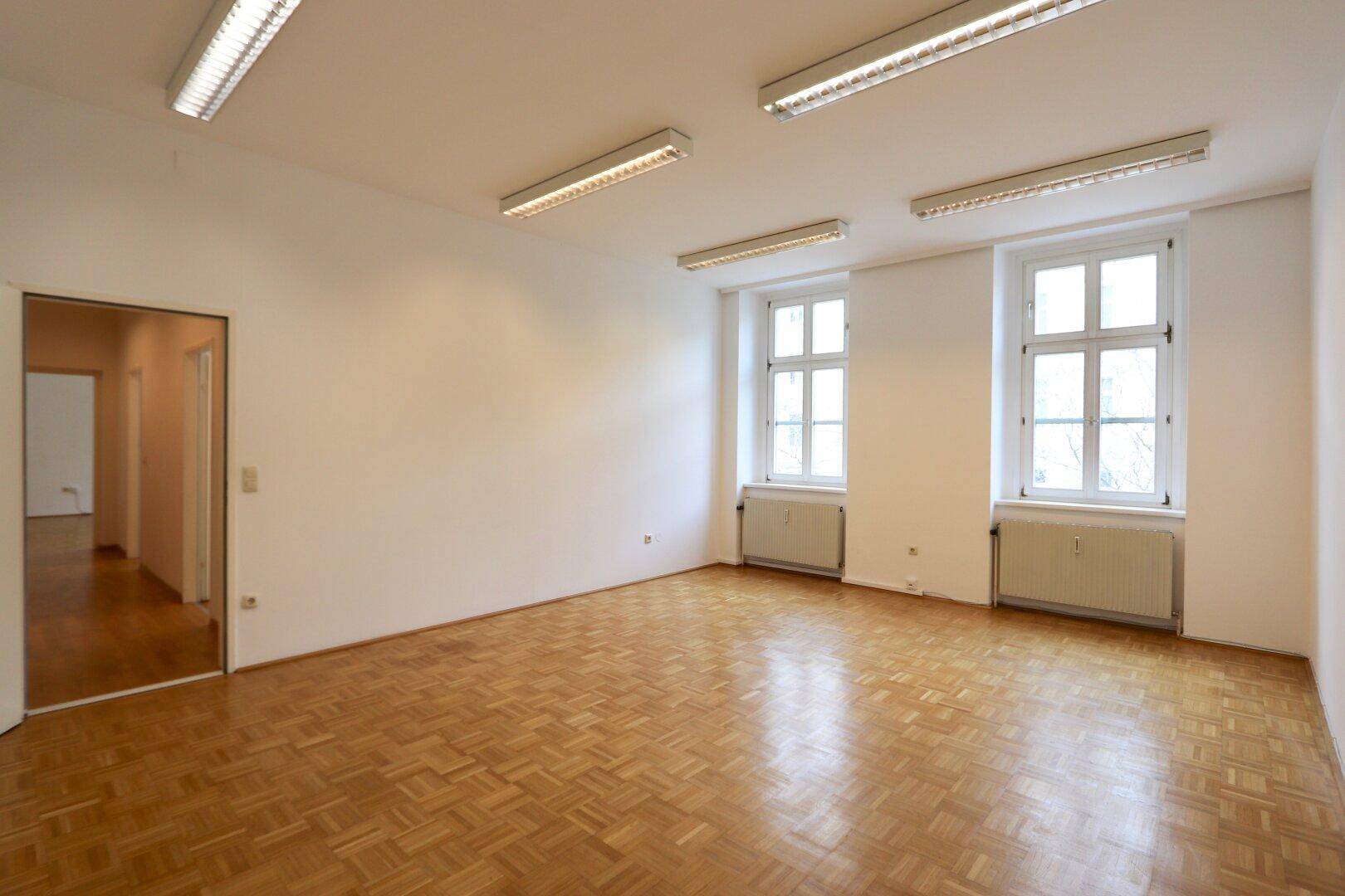 Geräumiger, heller Büroraum mit Parkettboden, zwei Fenstern und Deckenleuchten.