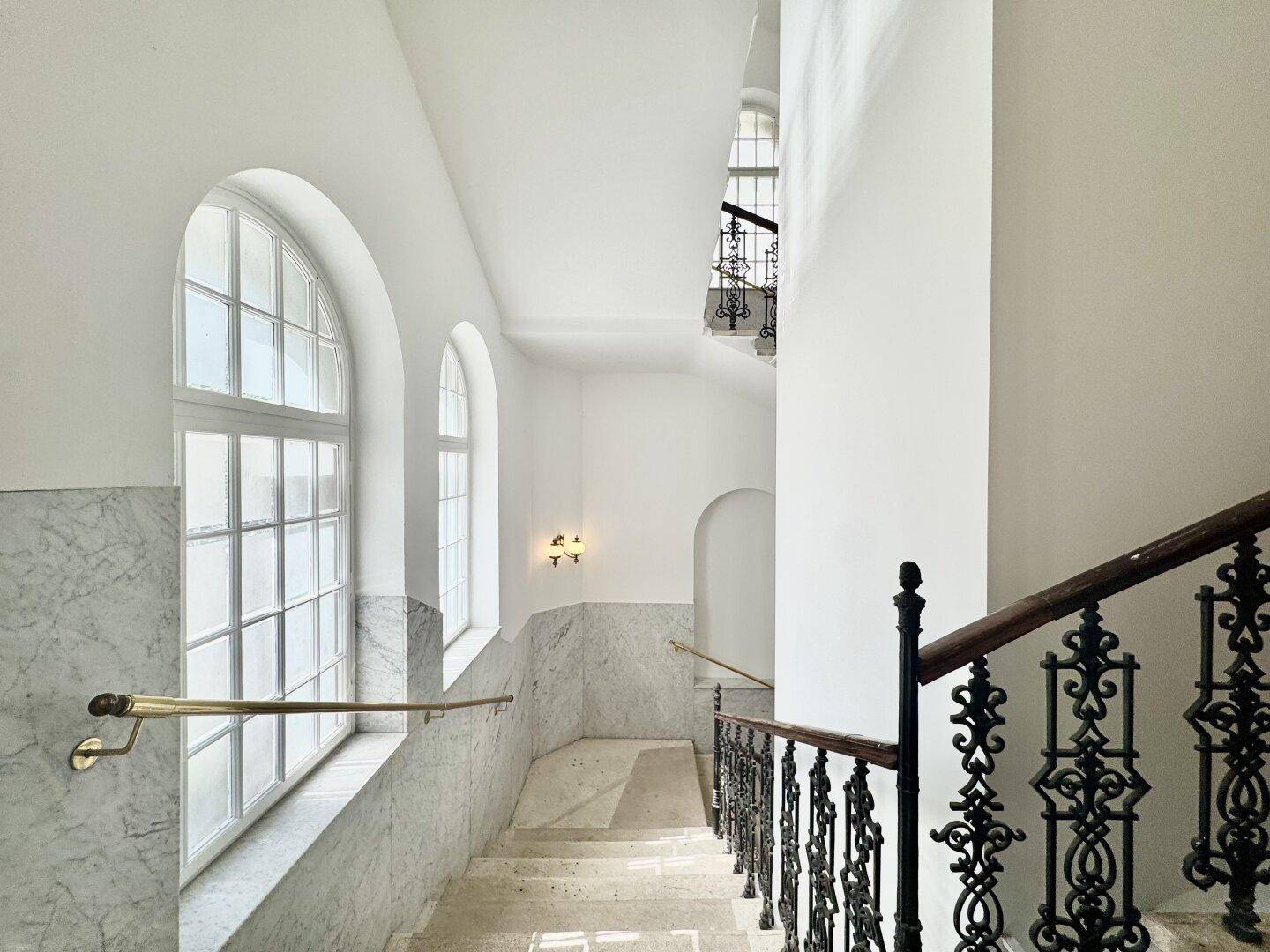 Helles Treppenhaus mit Marmorverkleidung, schmiedeeisernem Geländer und eleganten Bogenfenstern.