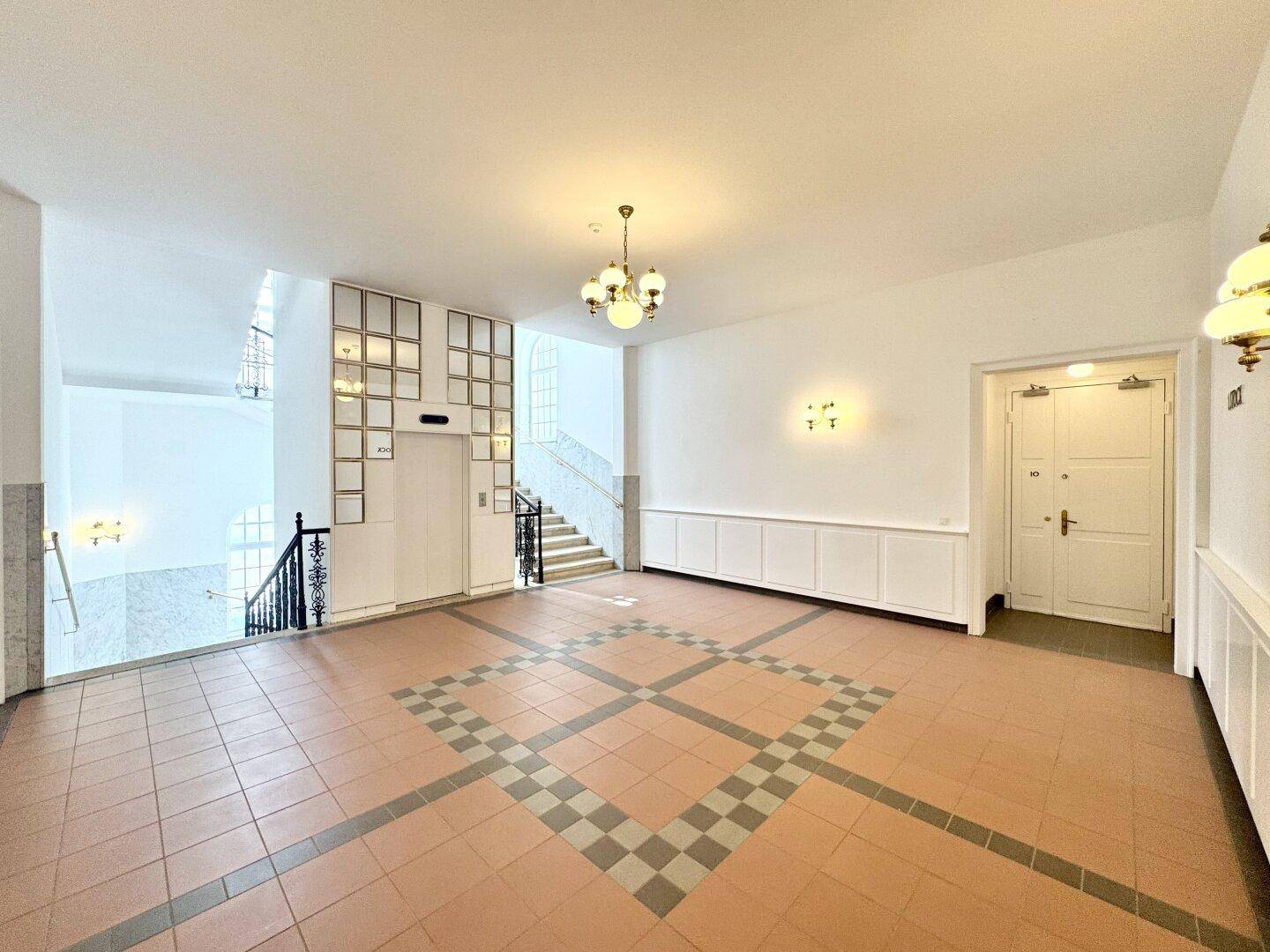 Geräumige Eingangshalle mit Fliesenboden, Aufzug, Treppenhaus und einem klassischen Kronleuchter.