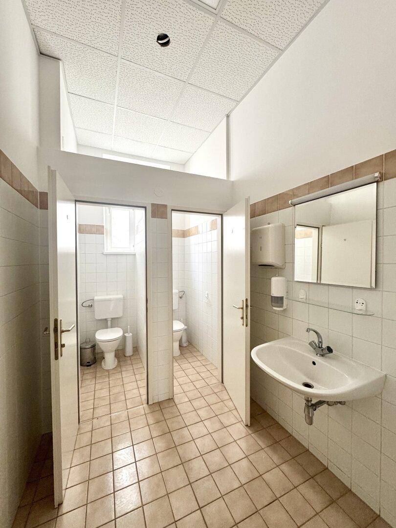 Mehrere Toilettenkabinen in einem öffentlichen Waschraum mit Fliesenboden und Waschbecken.