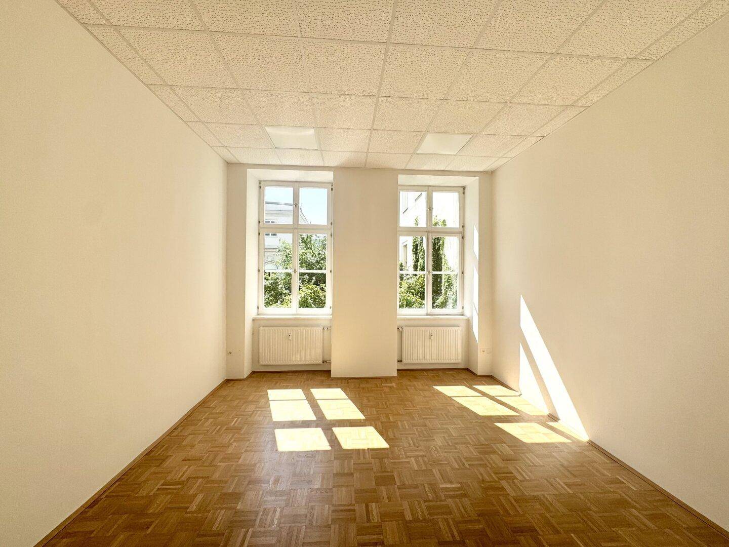 Heller Raum mit Holzboden und zwei Fenstern, durch die Sonnenlicht einfällt.