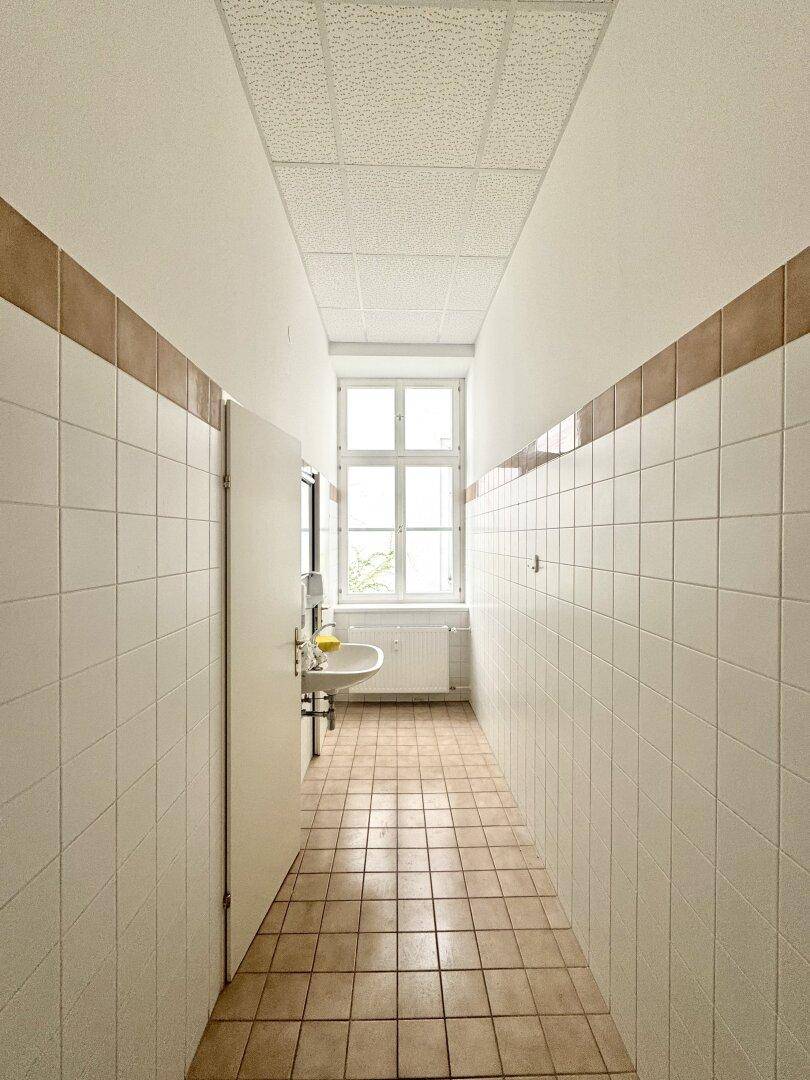 Langer Flur in einem Waschraum mit Fliesenboden, Waschbecken und Fenster.