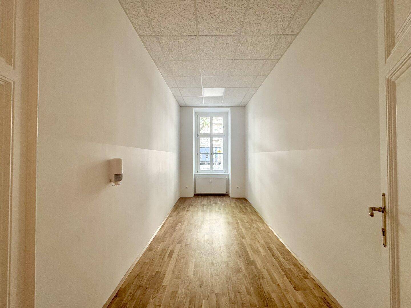Langer, heller Raum mit Holzboden und einem Fenster am Ende, ideal für ein Großraumbüro.