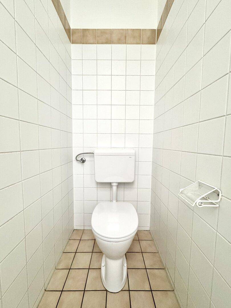 Schmale Toilette mit weißen Fliesen an den Wänden und einem hellen Fliesenboden.