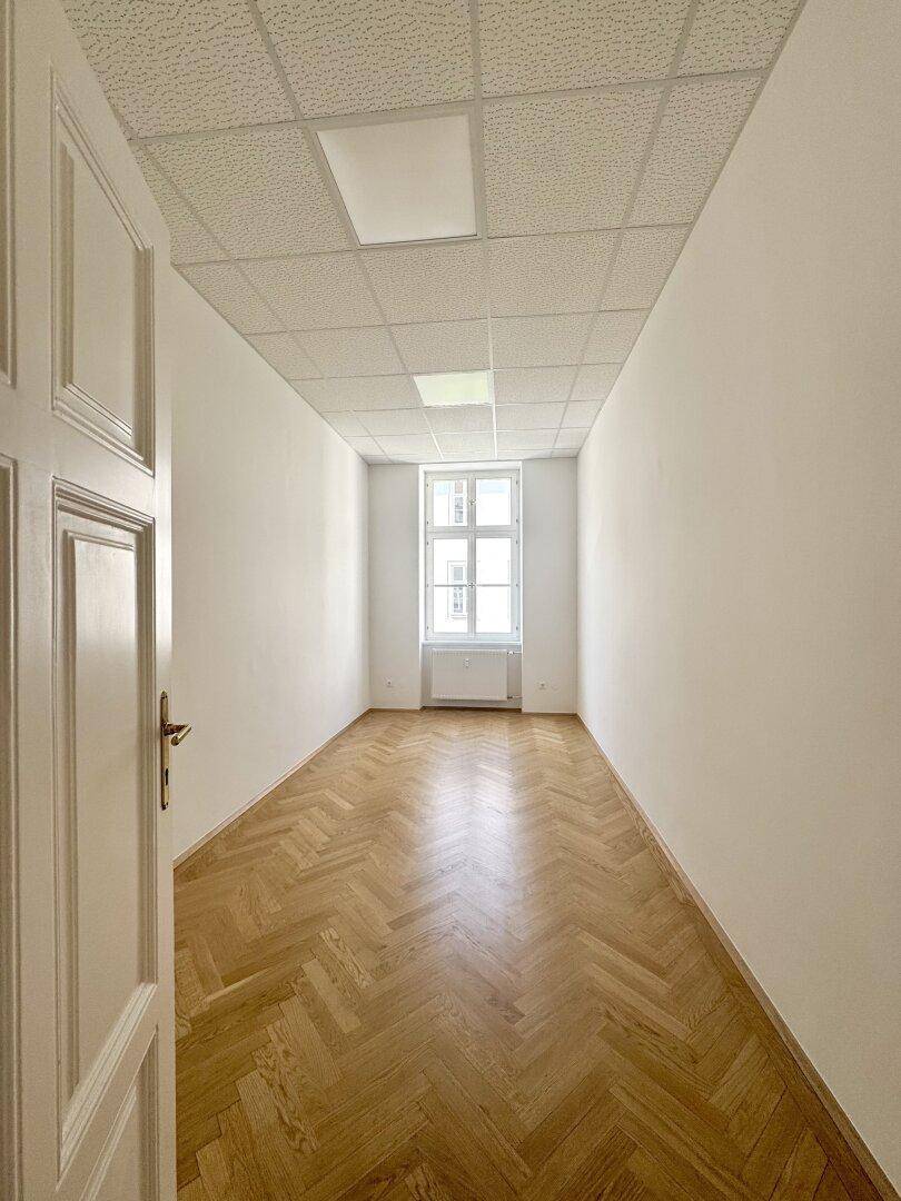 Heller, leerer Raum mit Fischgrätparkett, weißen Wänden und einem Fenster am Ende.