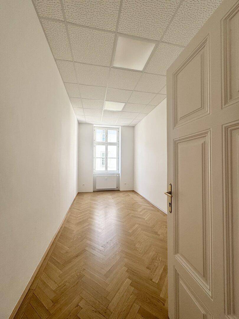 Heller, leerer Raum mit Fischgrätparkett, weißen Wänden und einem Fenster am Ende.