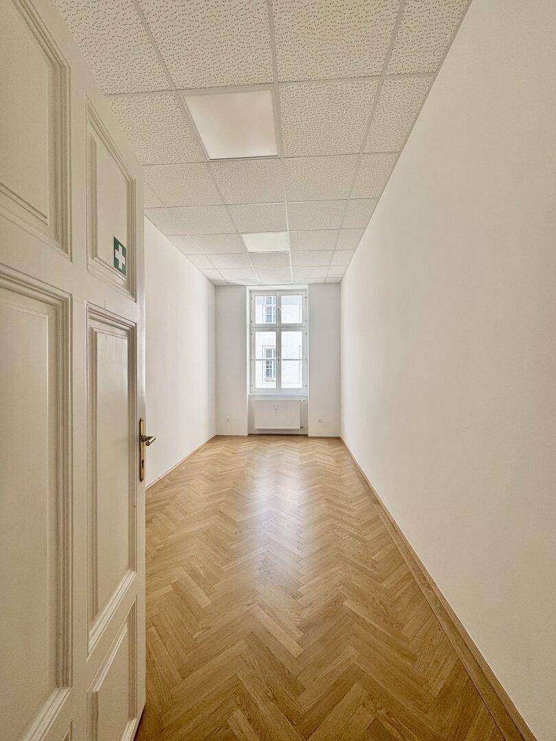 Langer, leerer Raum mit Fischgrätparkett, weißen Wänden und einem Fenster am Ende.