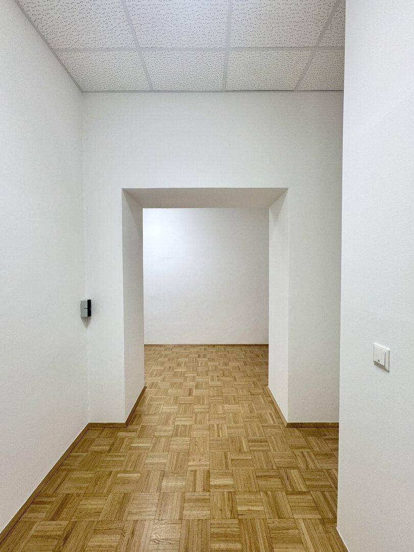 Heller Raum mit Parkettboden und einem offenen Durchgang zu einem weiteren leeren Bereich.
