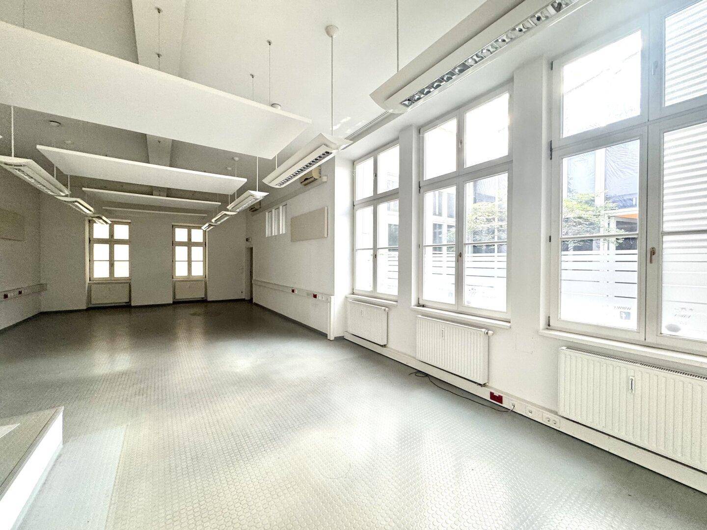 Großer, leerer Gewerberaum mit hohen Fenstern, Akustikplatten an der Decke und viel Tageslicht.