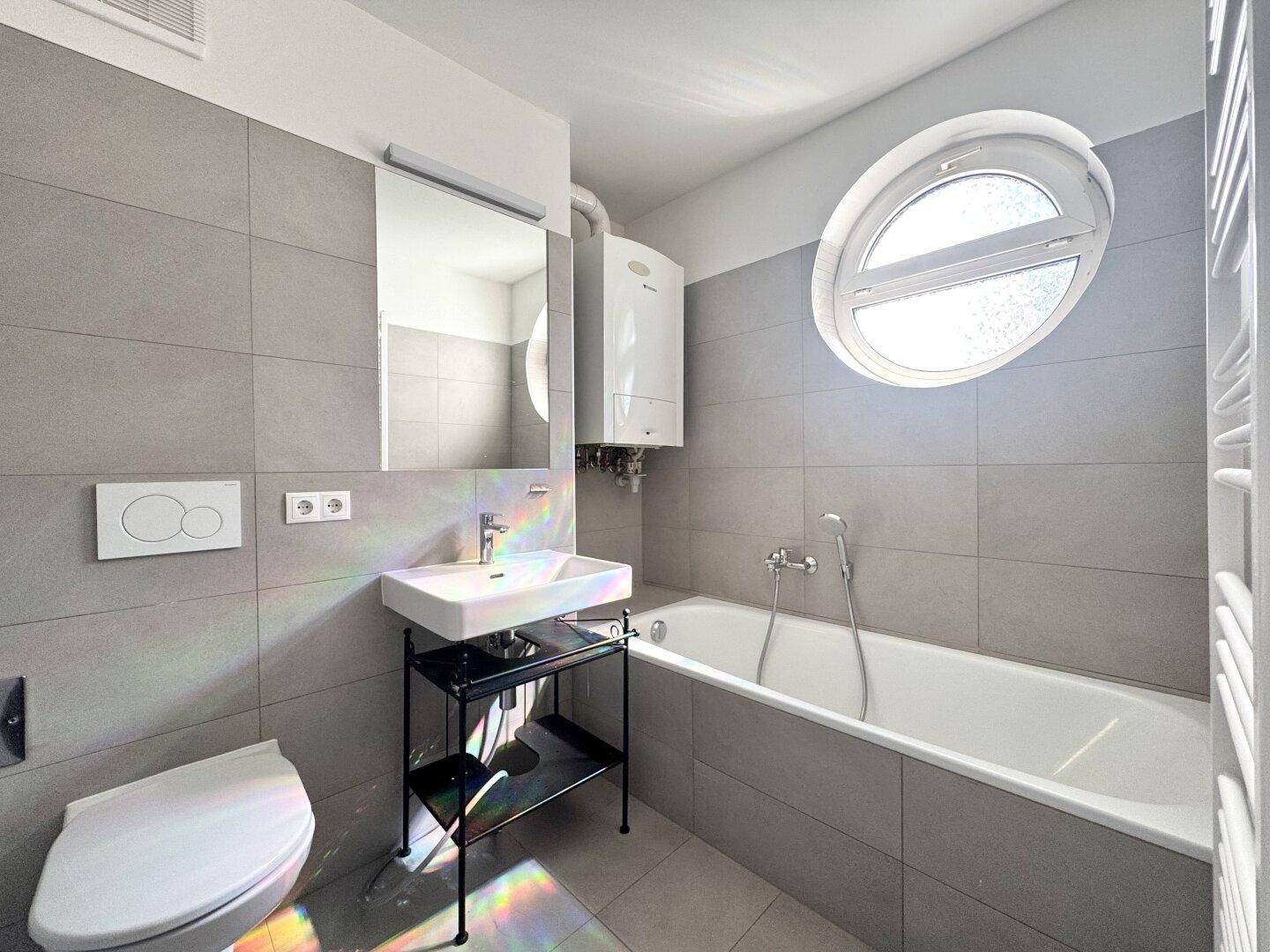 Modernes Badezimmer mit grauen Fliesen, Badewanne und einem einzigartigen runden Fenster.