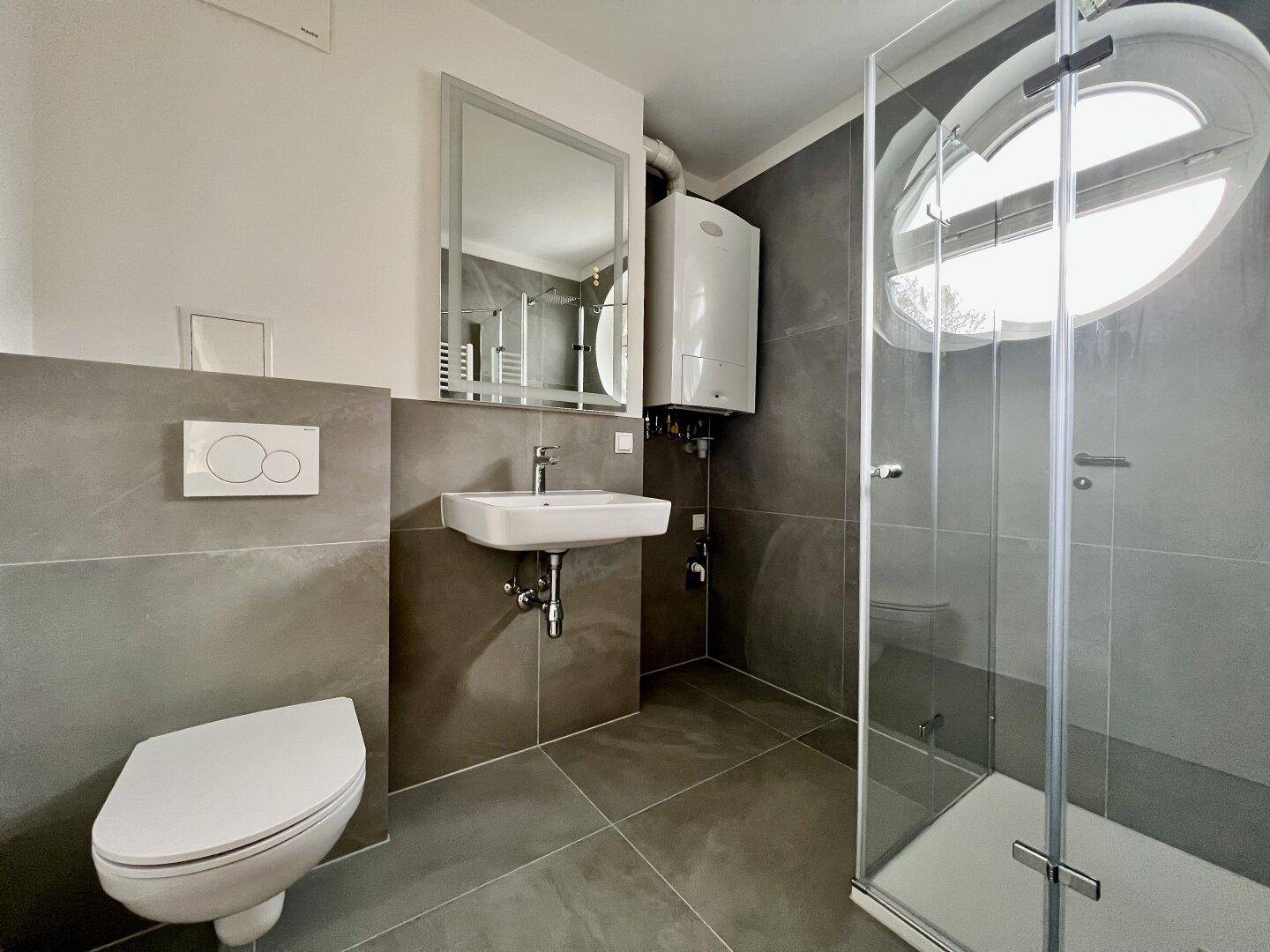 Modernes Badezimmer mit grauen Fliesen, wandhängendem WC, Waschbecken und Duschkabine mit Rundfenster.