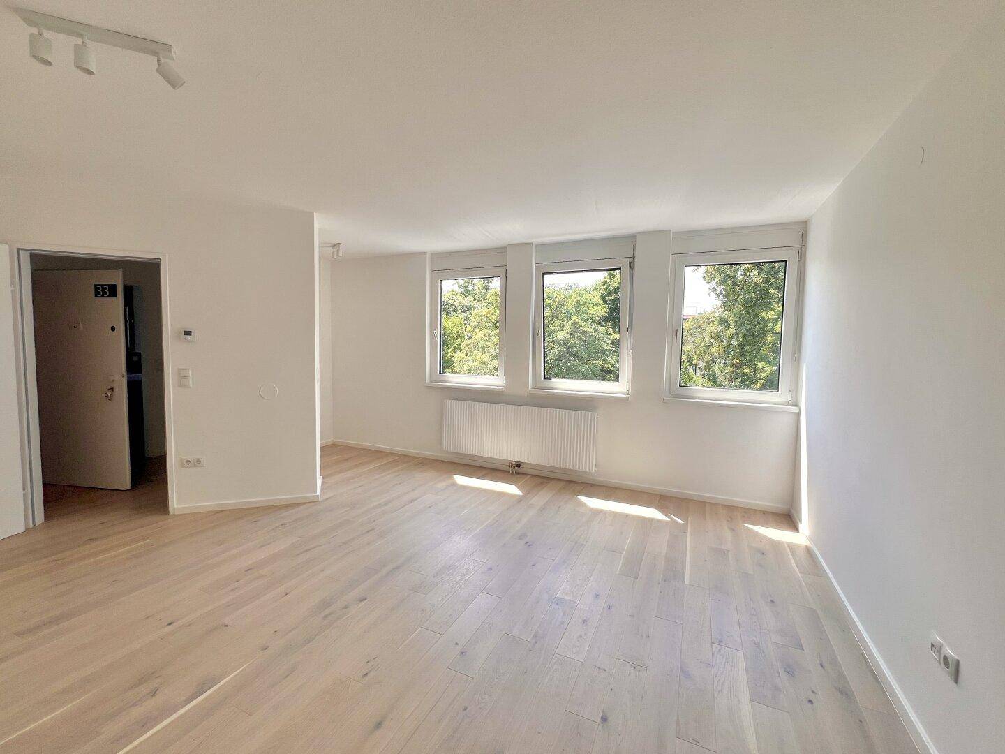Heller, leerer Raum mit hellem Holzparkett und Blick ins Grüne durch drei große Fenster.