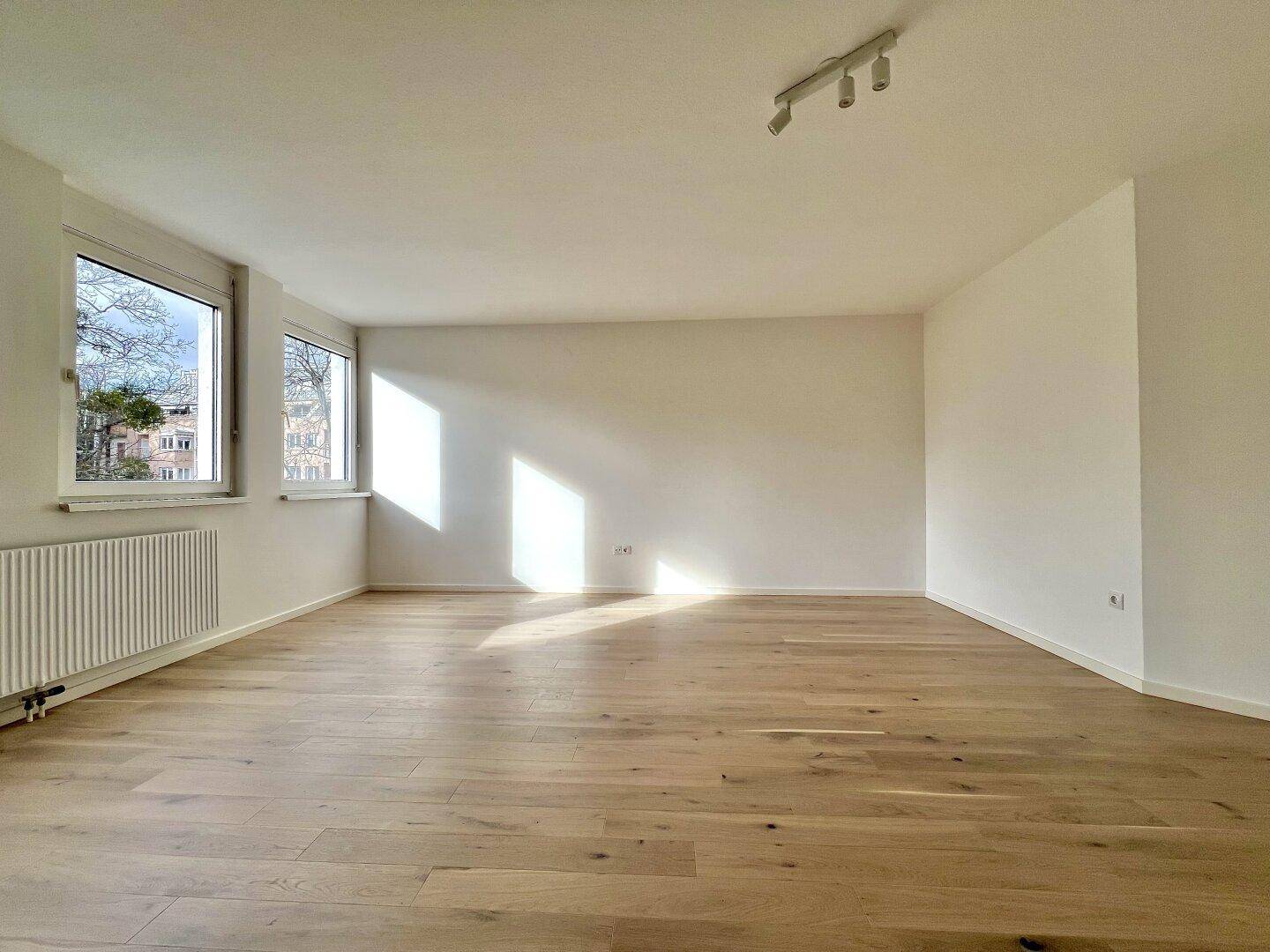 Lichtdurchfluteter Raum mit hellem Holzparkett, weißen Wänden und zwei Fenstern mit Blick nach draußen.