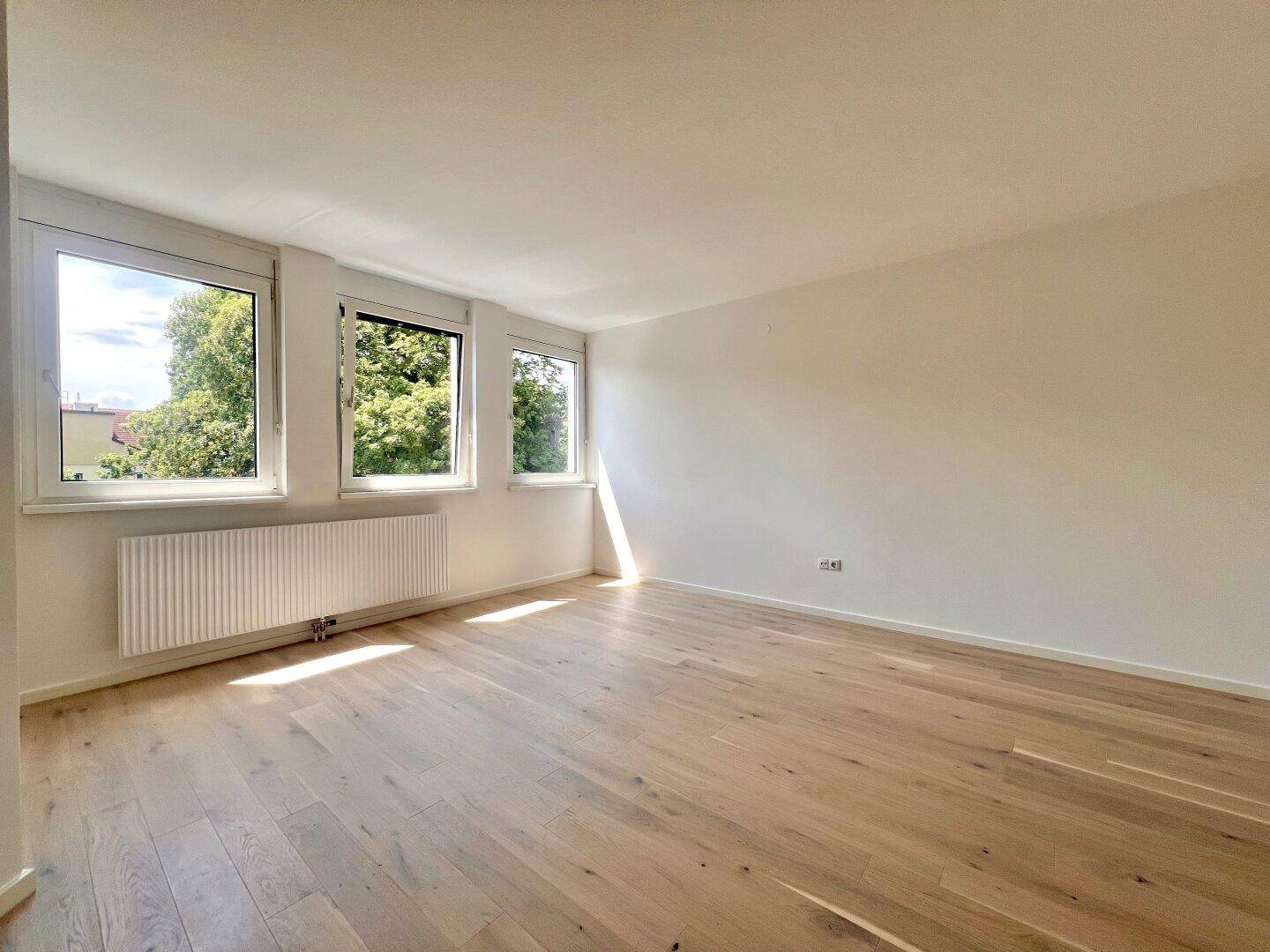 Heller, leerer Raum mit hellem Holzparkett und Blick ins Grüne durch drei große Fenster.