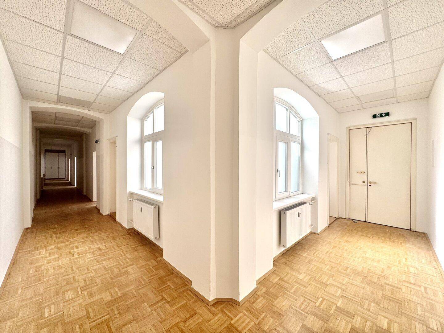 Langer, heller Flur mit Parkettboden, hohen Fenstern und einer Decke mit Rasterplatten.