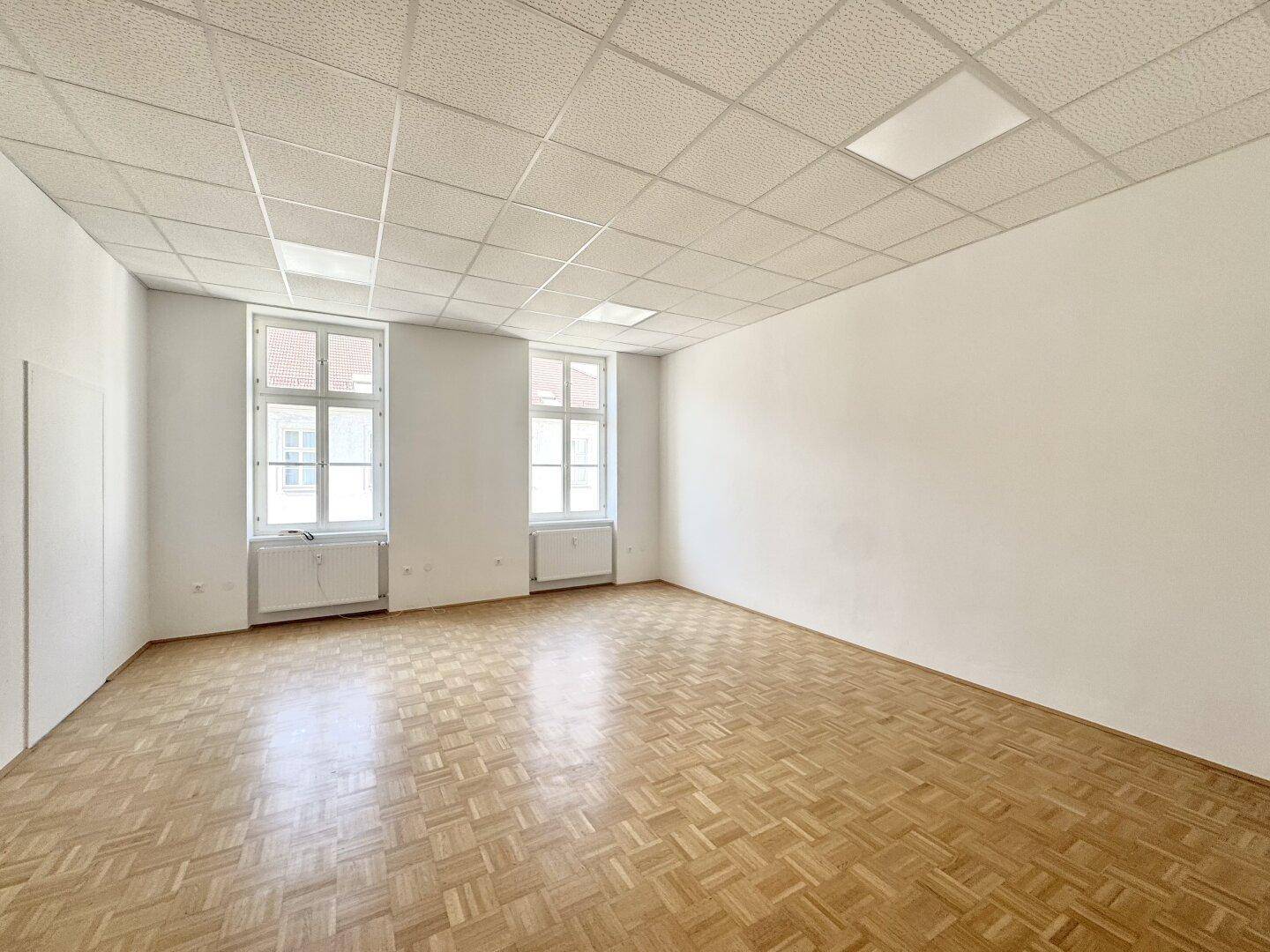 Heller und leerer Raum mit Parkettboden, weißen Wänden und zwei großen Fenstern.