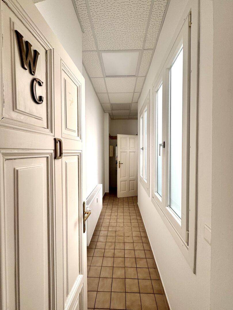 Schmaler Flur mit Fliesenboden, Fenstern und einer Tür, die zu einer Toilette führt.