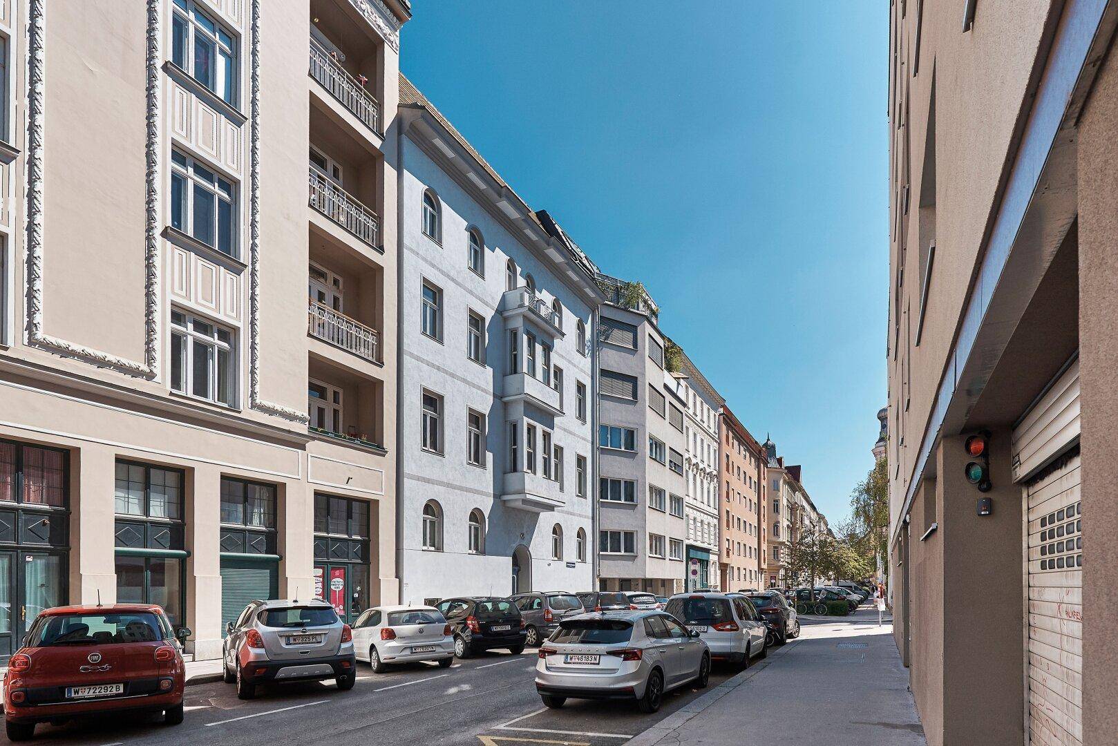 Eine belebte Stadtstraße mit geparkten Autos und historischen Gebäuden unter blauem Himmel.