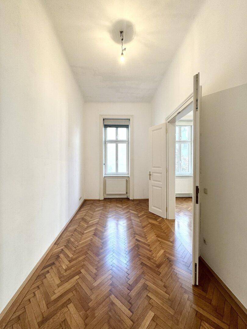 Langes, helles Zimmer mit Fischgrätparkettboden und einem Fenster, das in einen weiteren Raum führt.