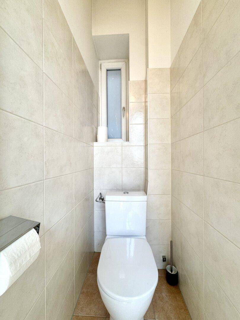Separate Toilette mit Wand-WC und kleinem Fenster, vollständig mit hellen Fliesen verkleidet.