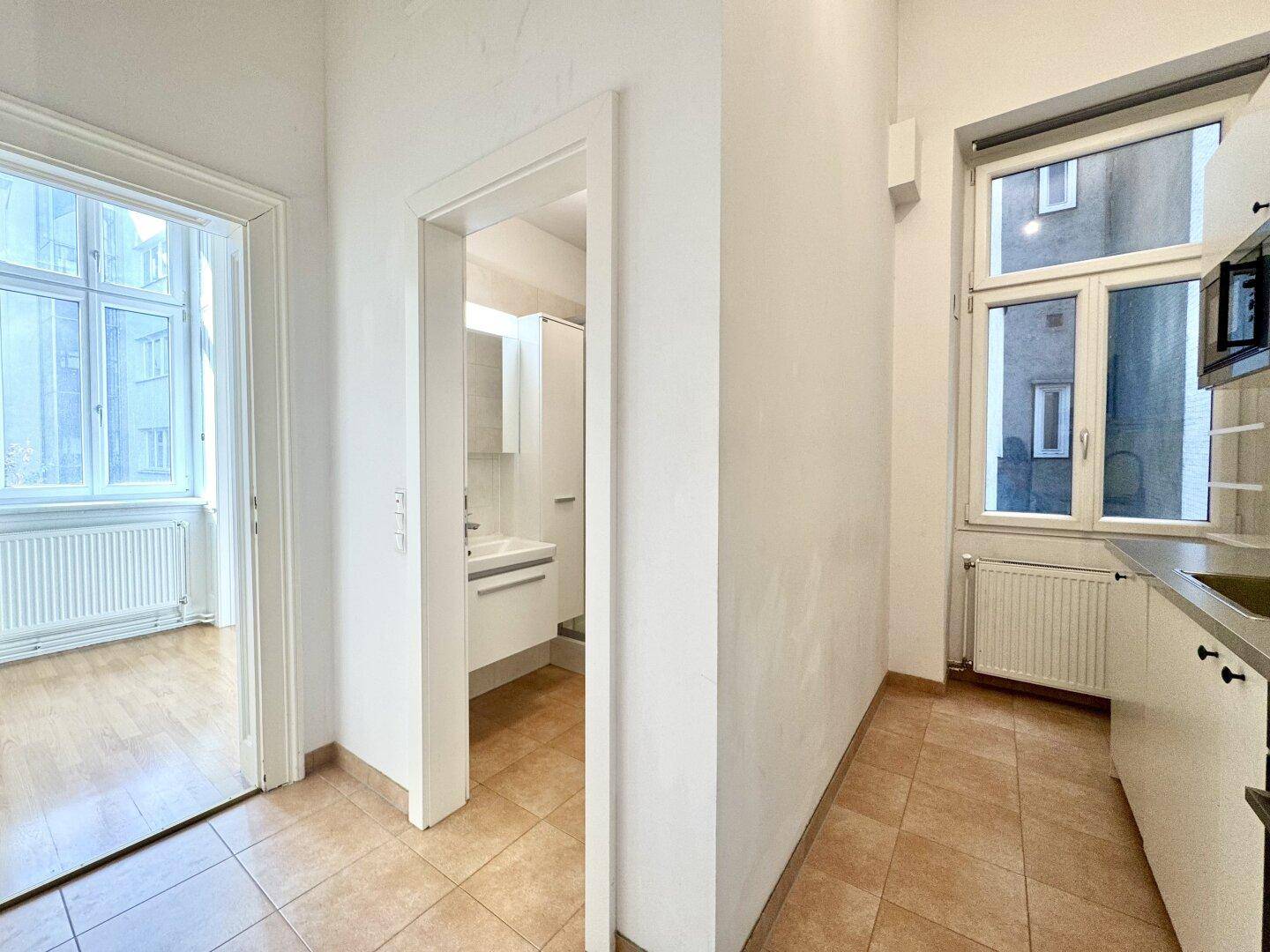 Flur mit Fliesenboden, der zu einer Küche und einem Badezimmer führt, mit Blick auf einen Innenhof.
