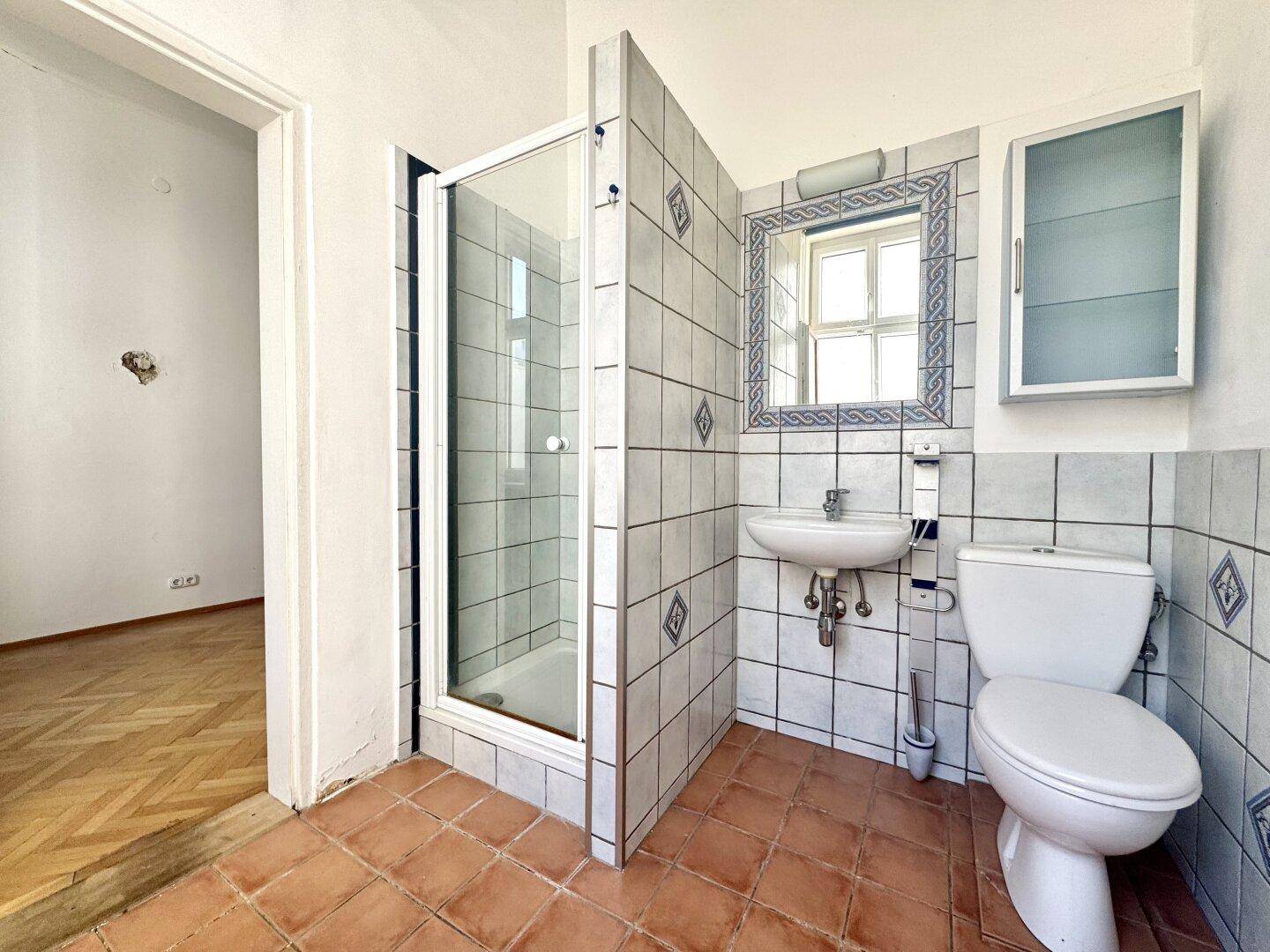 Kleines Badezimmer mit Duschkabine, WC und Terrakotta-Fliesen, angrenzend an ein Zimmer.