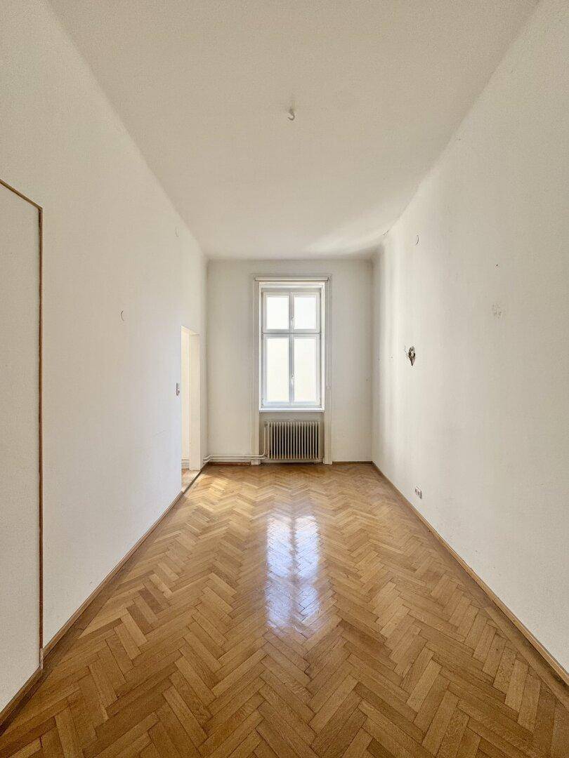 Helles, langes Zimmer mit Fischgrätparkett und einem Fenster, ideal als Schlafzimmer.