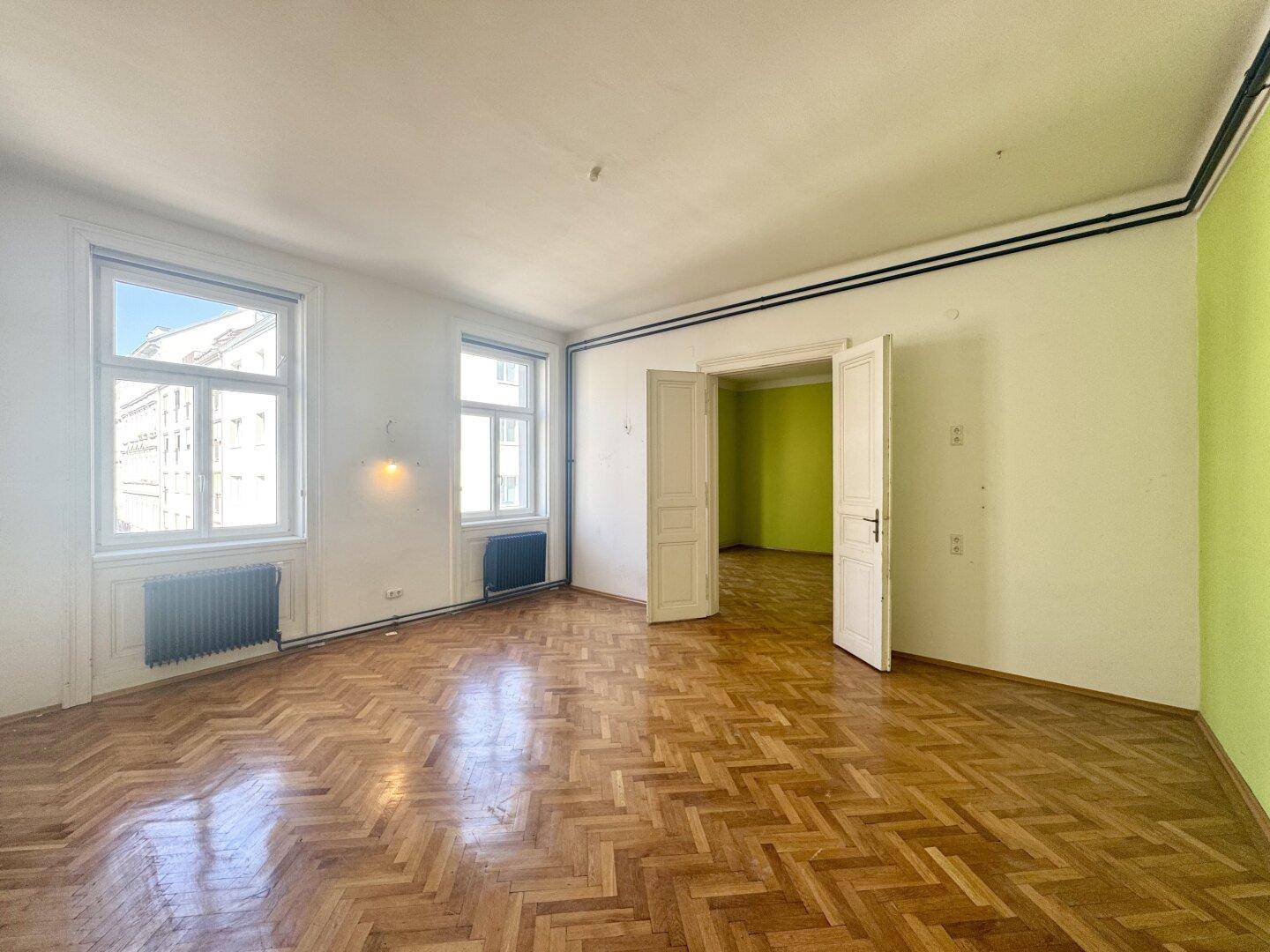 Helles Zimmer mit Fischgrätparkett und hohen Fenstern, teilweise mit grünen Wänden.