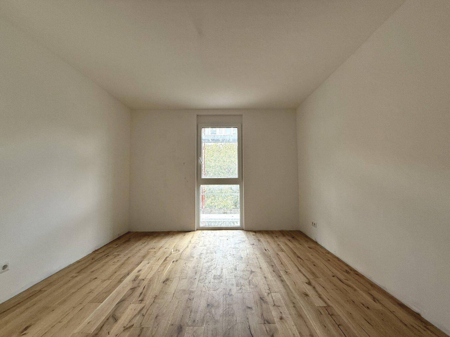 Leeres Zimmer mit hellem Holzboden und einem hohen Fenster für viel Tageslicht.