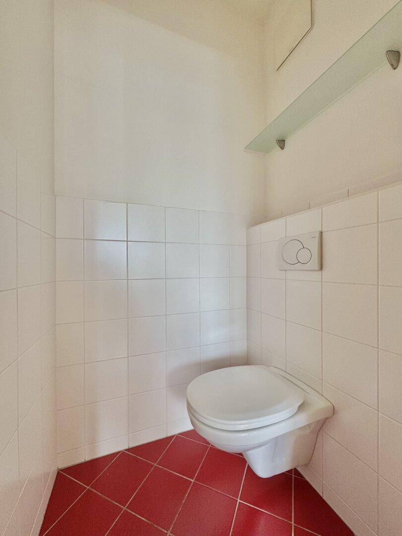 Separates WC mit wandhängender Toilette und roten Bodenfliesen.