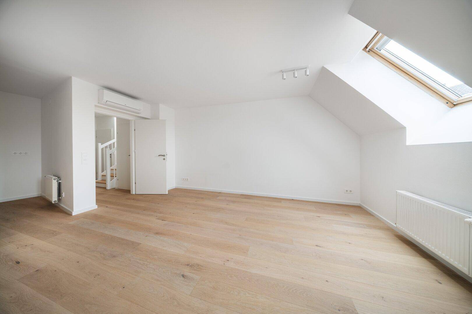 Helles, leeres Zimmer mit Dachschräge, Holzboden und Klimaanlage.