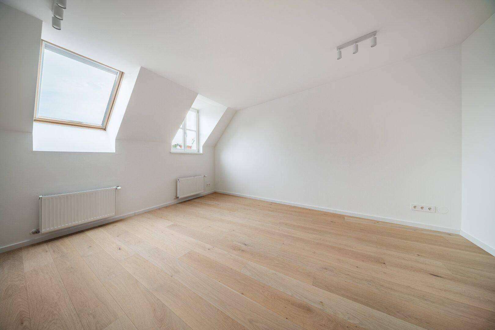Lichtdurchflutetes Zimmer mit Dachschrägen, Holzboden und zwei Dachfenstern.