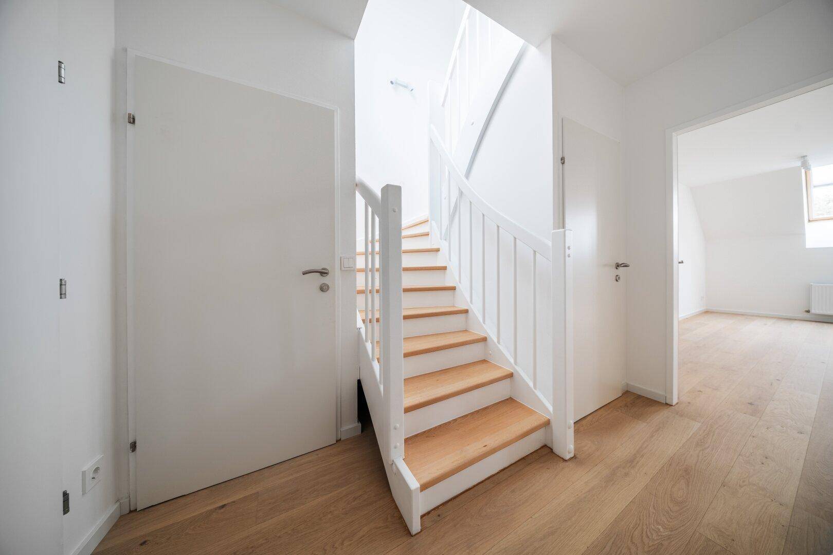 Heller Flur mit Holztreppe und weißen Wänden, der zu den oberen Etagen führt.