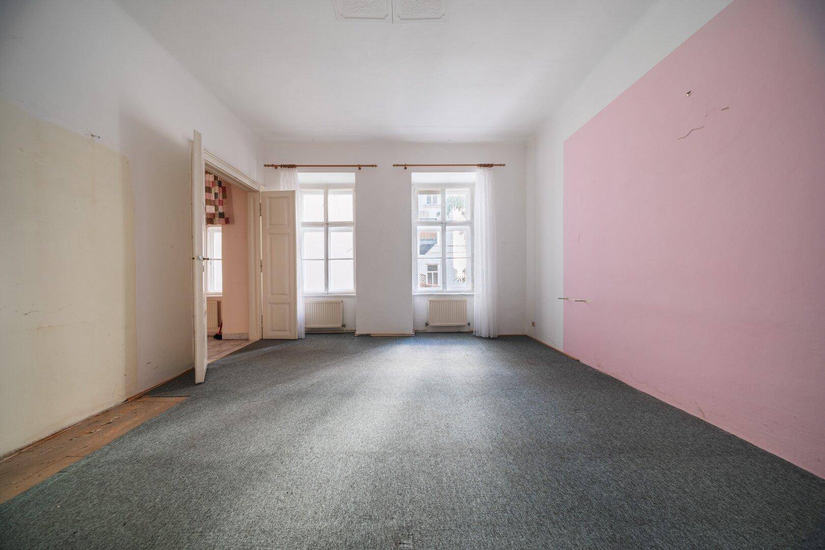 Leerer Raum mit grauem Teppichboden und einer rosa gestrichenen Wand, bereit zur Renovierung.