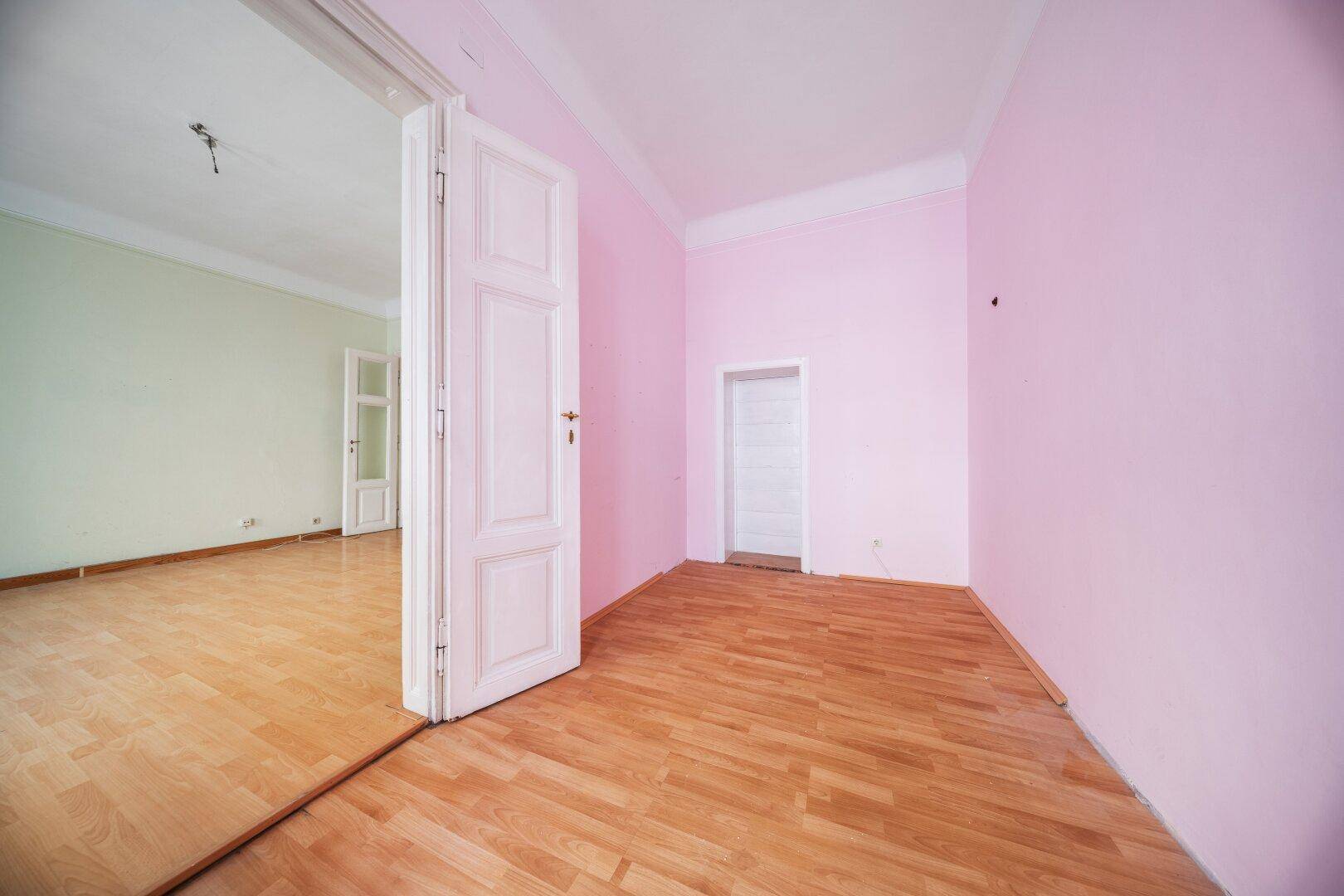 Leerer Raum mit rosa Wänden und hellem Laminatboden, verbunden mit einem weiteren Zimmer.