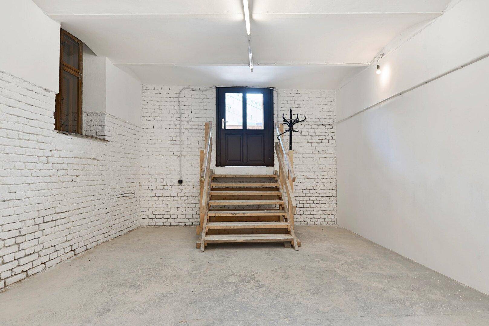 Heller Kellerraum mit weiß gestrichenen Ziegelwänden, Holztreppe und Zugang nach draußen.