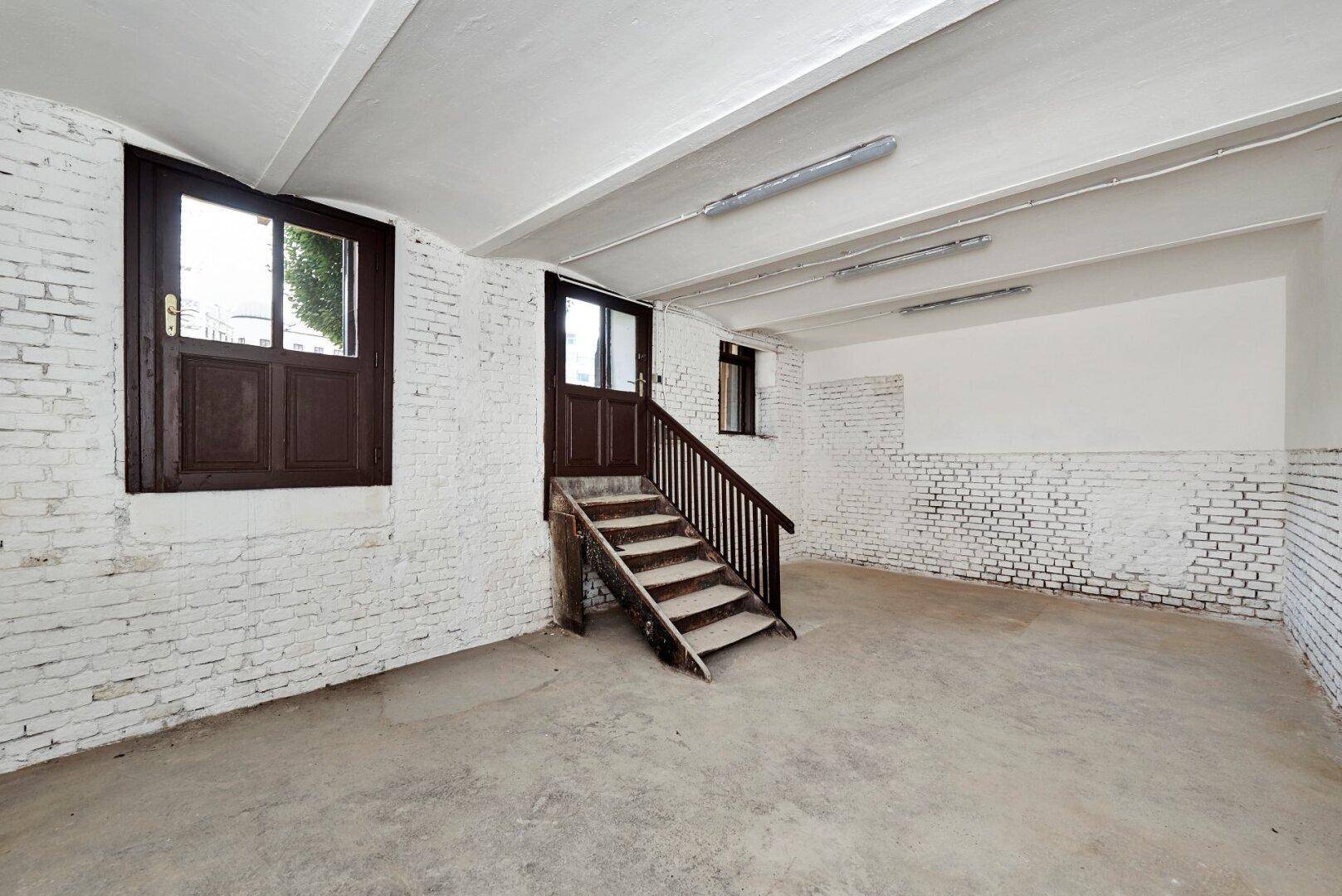 Geräumiger Kellerbereich mit weiß gestrichenen Ziegelwänden, Betonboden und Holztreppe.