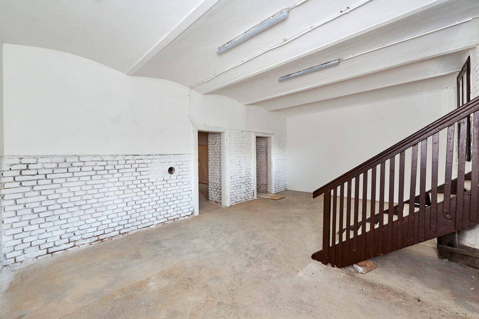 Weitläufiger Kellerraum mit weiß gestrichenen Ziegelwänden, Betonboden und Holztreppe.