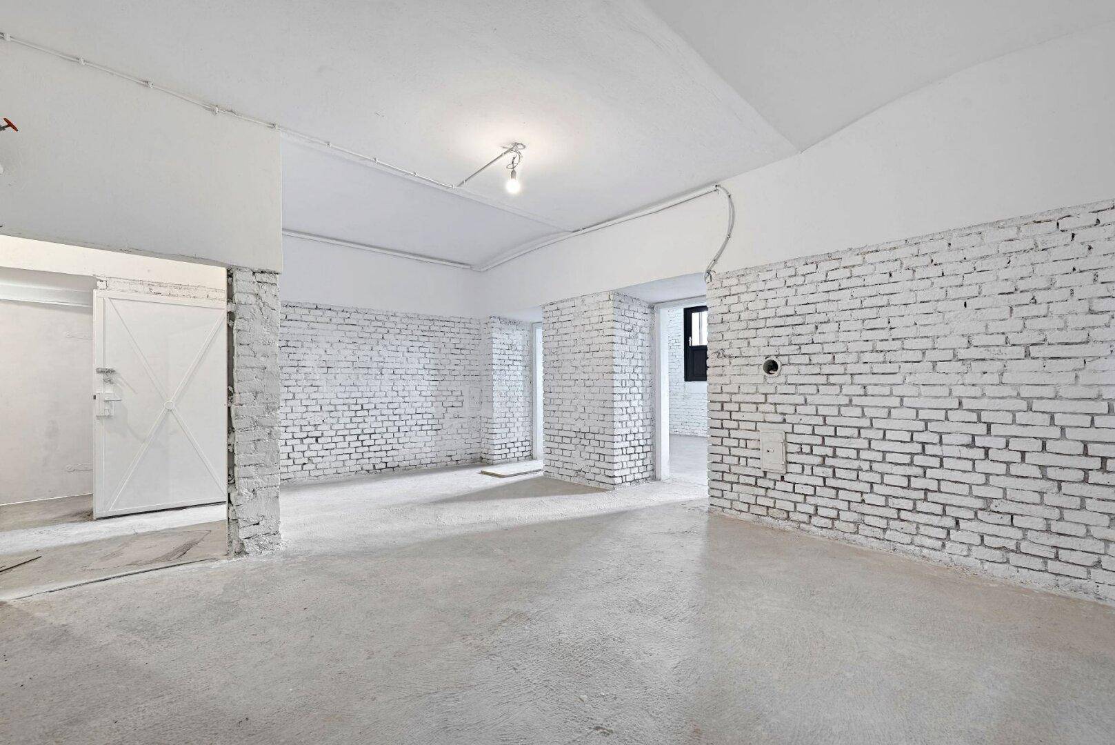 Großer Kellerraum mit weiß gestrichenen Ziegelwänden, Betonboden und mehreren Türöffnungen.