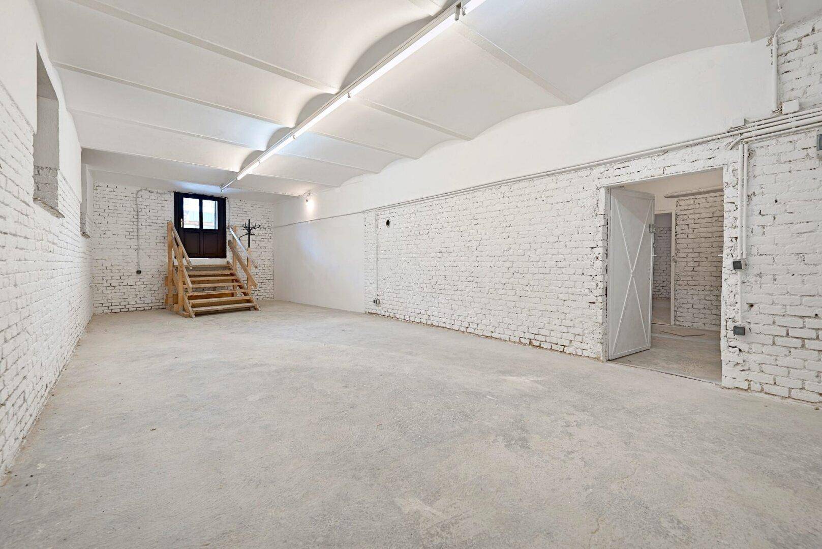 Geräumiger Kellerraum mit weiß gestrichenen Ziegelwänden, Betonboden und Holztreppe.