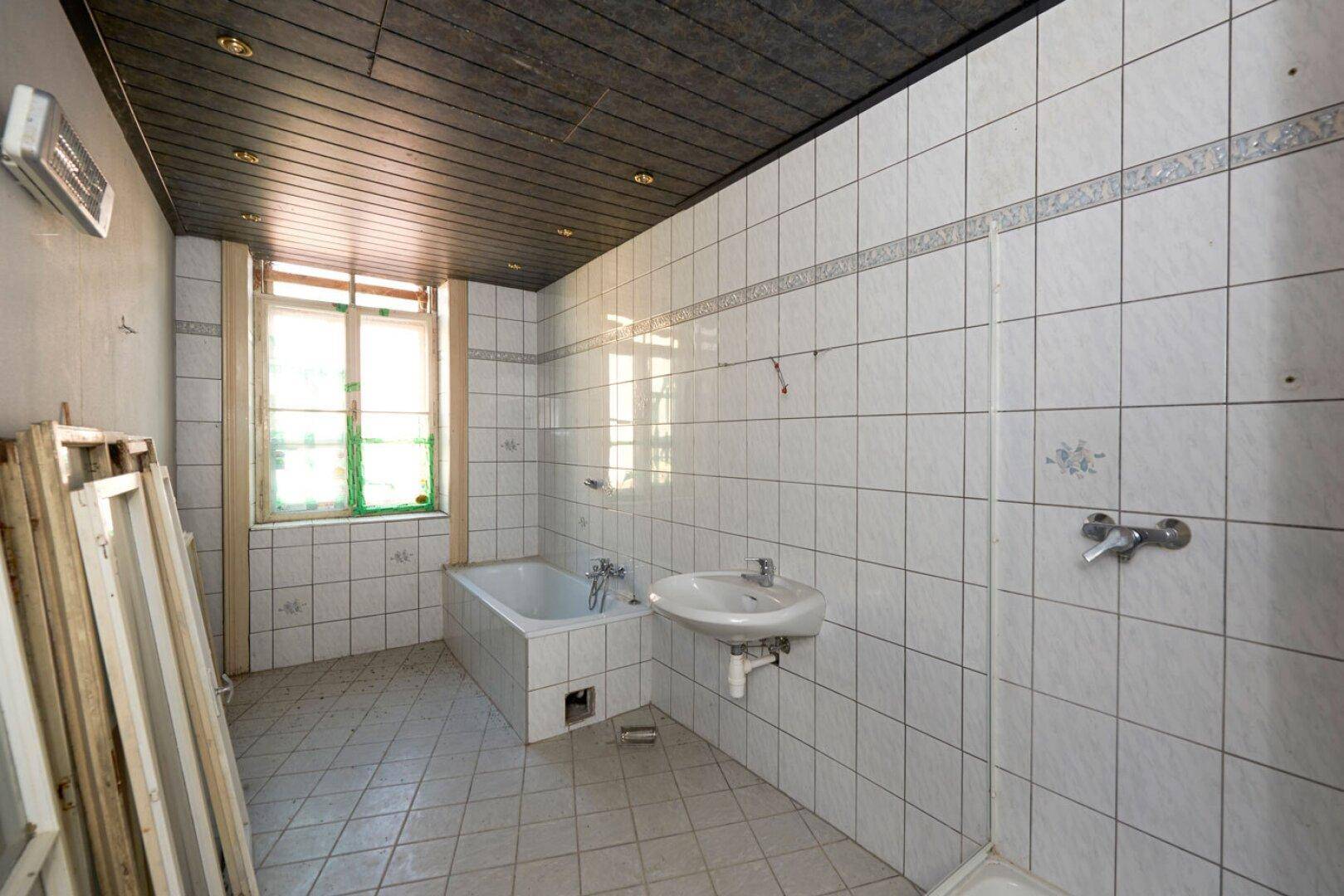 Badezimmer mit weißen Fliesen, Badewanne und Waschbecken, das eine Modernisierung erfordert.