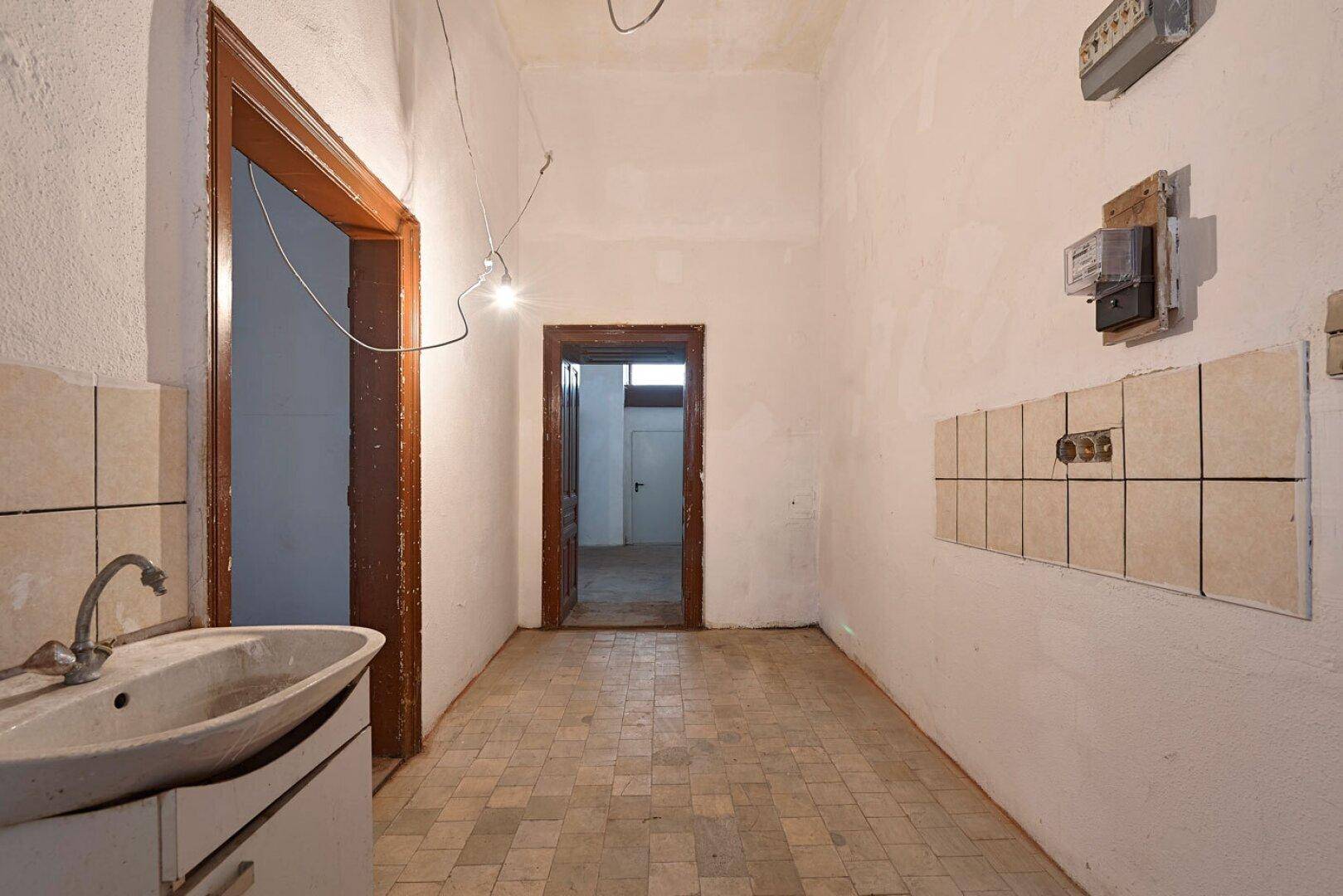 Langer, unrenovierter Küchenraum mit Fliesenboden, Waschbecken und sichtbaren Elektroinstallationen.