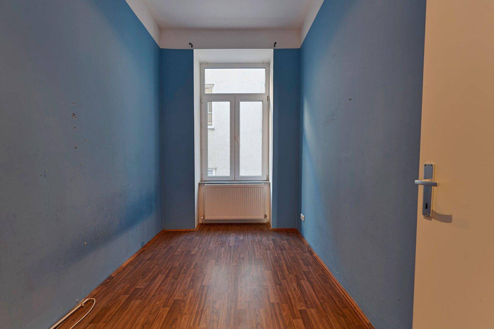 Kleines Zimmer mit blauen Wänden, Holzboden und einem Fenster mit Heizkörper.
