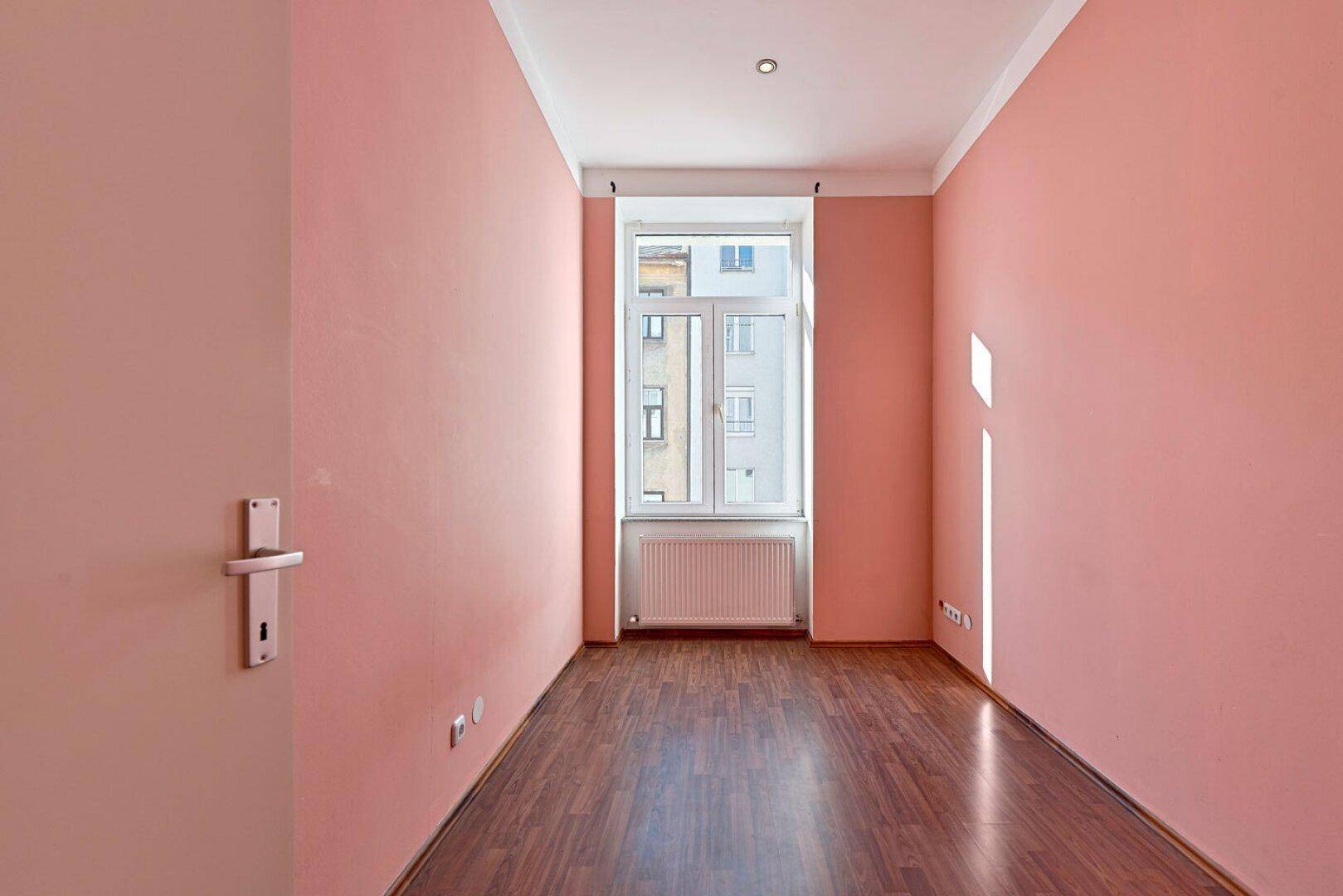 Schmaler Raum mit rosa Wänden, Holzboden und einem Fenster mit Heizkörper.